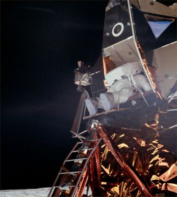AS 11-40-5863 (HD) - Buzz's going down the ladder
Buzz has his PLSS most of the way out of the hatch. His feet are near the lip of the porch. Neil has moved around to the south of the ladder a little bit. Note the near-side handrail and the triangular Commander's window above and to the right of the hatch. There is a reflection of the lunar surface in the window. To Buzz's left, we can see a set of thrusters and the plume deflector that protects the LM from the downward thruster exhaust. 
Parole chiave: LM
