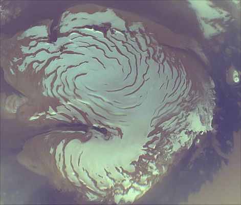 The "North Polar Cap" of Mars
During the last week of September and the first week or so of October, 2006, the Mars Reconnaissance Orbiter (MRO) scientific instruments were turned on to acquire test information leading up to full science operations to begin the first week of November 2006, following superior conjunction (superior conjunction is where a planet goes behind the sun as viewed from the Earth). Since it is very difficult to communicate with a spacecraft when it is close to the Sun as seen from Earth, this checkout of the instruments was crucial to being ready for the Primary Science Phase (PSP) of the mission. 

Throughout the Transition Phase of operations, the Mars Color Imager (MARCI) acquired terminator (transition between nighttime and daytime) to terminator swaths of color images on every dayside orbit, as the spacecraft moved northward in its orbit. The south polar region was deep in winter shadow, but the north polar region was illuminated the entire martian day. During the primary mission, these swaths will be assembled into global maps that portray the state of the martian atmosphere -- its weather -- as seen every day and at every place at about 3 PM local solar time. After the Transition Phase was completed, most of the instruments were turned off, but the Mars Climate Sounder and MARCI have been left on. Their data will be recorded and played back to Earth following the communications blackout associated with conjunction and just prior to the start of the PSP. 

Combined with Mars Global Surveryor (MGS) Mars Orbiter Camera (MOC) wide angle image mosaics taken at 2 PM local solar time, the MARCI maps will be used to track motions of clouds. 

The image shown above is a composite mosaic of four polar views of Mars, taken at midnight, 6 AM, noon, and 6 PM local martian time. This is possible because during summer the sun is always shining in the polar region. It shows the mostly water ice perennial cap (white area), sitting atop the north polar layered materials (light tan immediately adjacent to the ice), and the dark circumpolar dunes. This view shows the region poleward of about 72 degrees north latitude. The data were acquired at about 900 meters per pixel. Three channels are shown here (425 nm, 550 nm, and 600 nm). 

Parole chiave: Mars from orbit - North Polar Cap