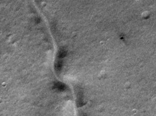 Dry Riverbed and a possible Skylight (or an EXTREMELY fresh Impact Crater) between Coracis Fossae and Bosporos Planum (EDM - credits: Lunexit)
Il rilievo ambiguo è sostanzialmente indefinibile a questa risoluzione, ma la sua colorazione "pitch black" e l'apparente assenza di rim ci fa pensare ad un Pozzo Verticale da Collasso.
Le "sbavature" scure che caratterizzano l'intero versante Nord della Surface Feature e, in parte, il lato Est-Sud/Est (senza interessamento della sua zona Ovest ed Ovest-Sud/Ovest), invece, potrebbero anche far pensare ad un impatto il quale, oltre che essere occorso assai di recente, potrebbe - se non altro parzialmente - essere ricondotto alla Classe dei "Mid-Air Impact" (ossia il bolide generatore del dettaglio si sarebbe, in questo caso, parzialmente disintegrato - esploso - in prossimità del suolo, mentre una sua parte avrebbe comunque raggiunto la superficie, così creando un cratere).

L'ipotesi del Vertical Collapse Pit, comunque, ci sembra preferibile (anche se una ragionevole spiegazione delle "sbavature" scure, in questo scenario, non siamo in grado di poterla fornire).
Parole chiave: Mars from orbit - Channel - Coracis Fossae - Bosporos Planum
